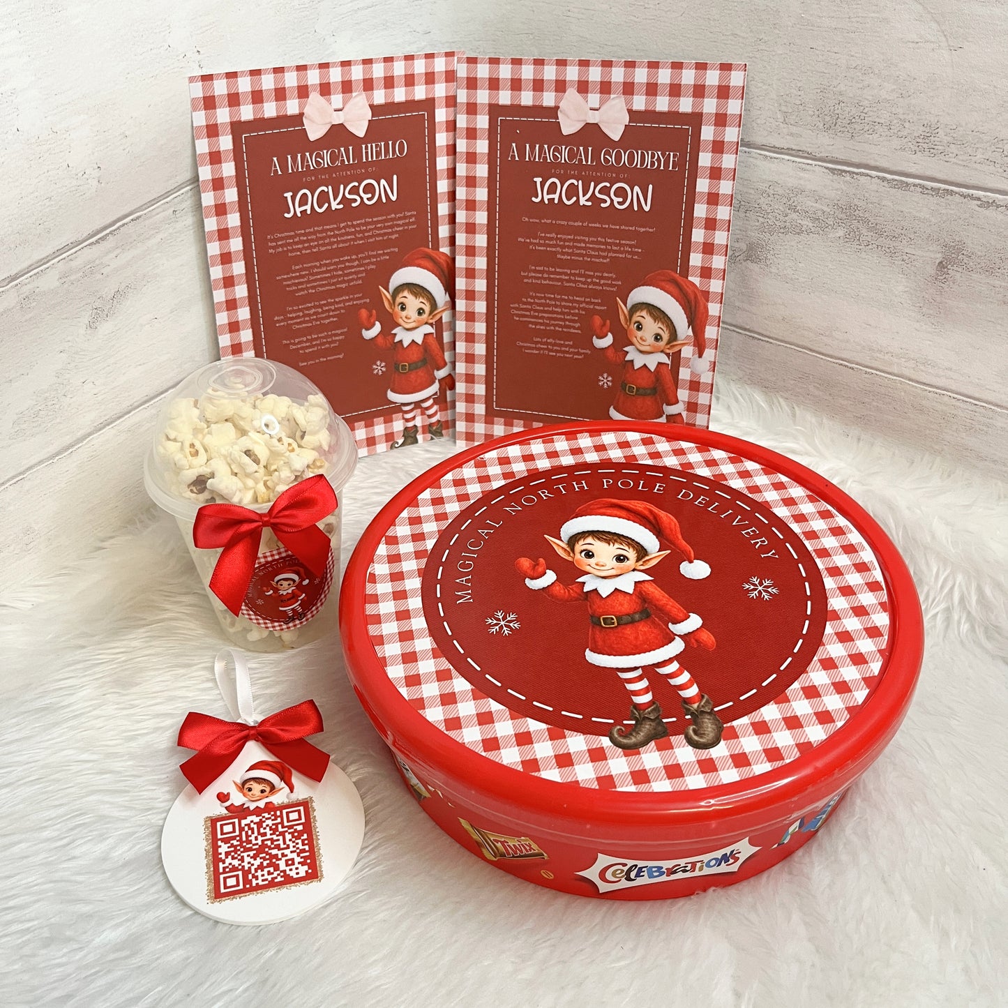 A Christmas-themed gift set featuring a round red and white gingham tin with an elf illustration on the lid, a small clear cup of popcorn tied with a red bow, a white ornament tag with a QR code and red bow, and two matching printed cards standing behind the tin.
