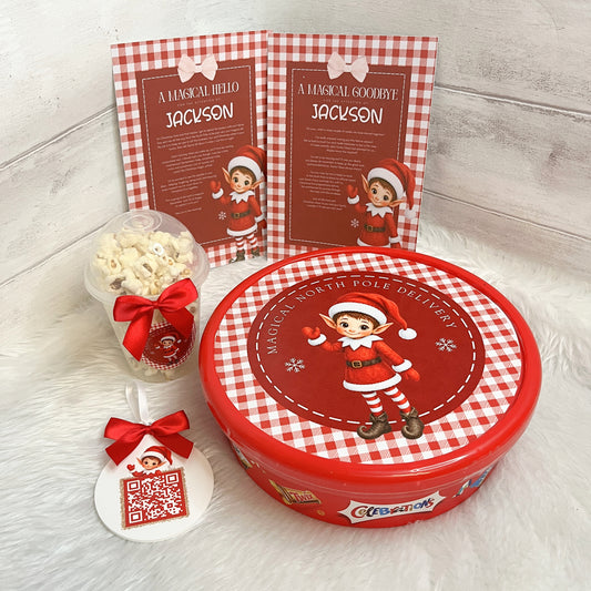 A Christmas-themed gift set featuring a round red and white gingham tin with an elf illustration on the lid, a small clear cup of popcorn tied with a red bow, a white ornament tag with a QR code and red bow, and two matching printed cards standing behind the tin.