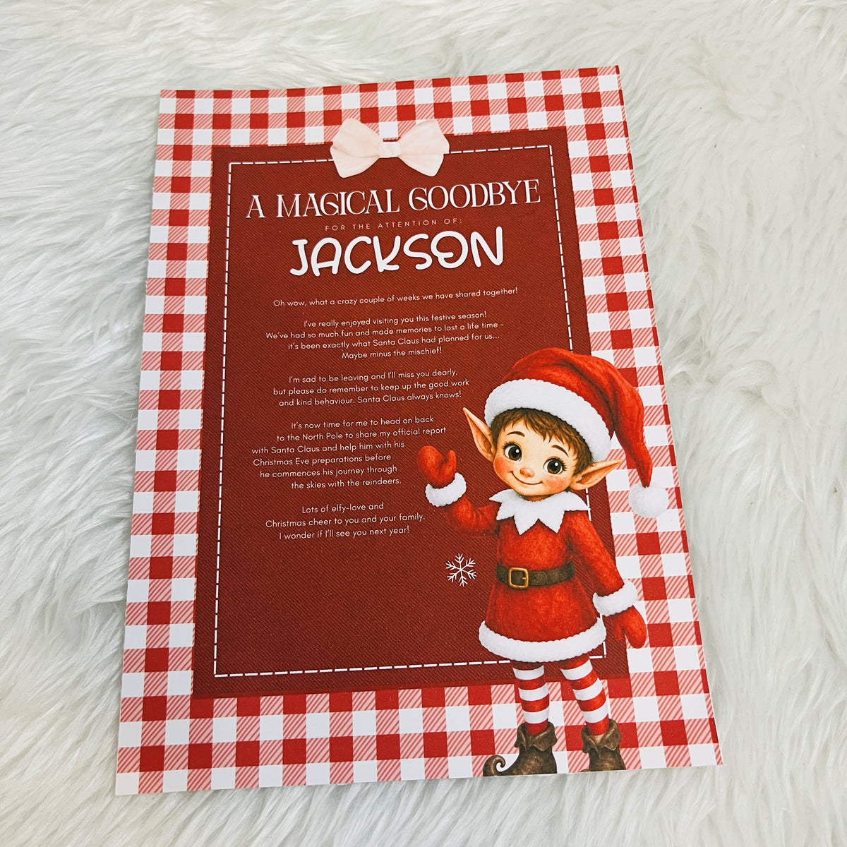 A personalized Christmas 'A Magical Goodbye' card featuring a red and white gingham border, a red central background with white text, a pink bow graphic at the top, and an elf illustration on the bottom right, displayed on a white faux fur surface.