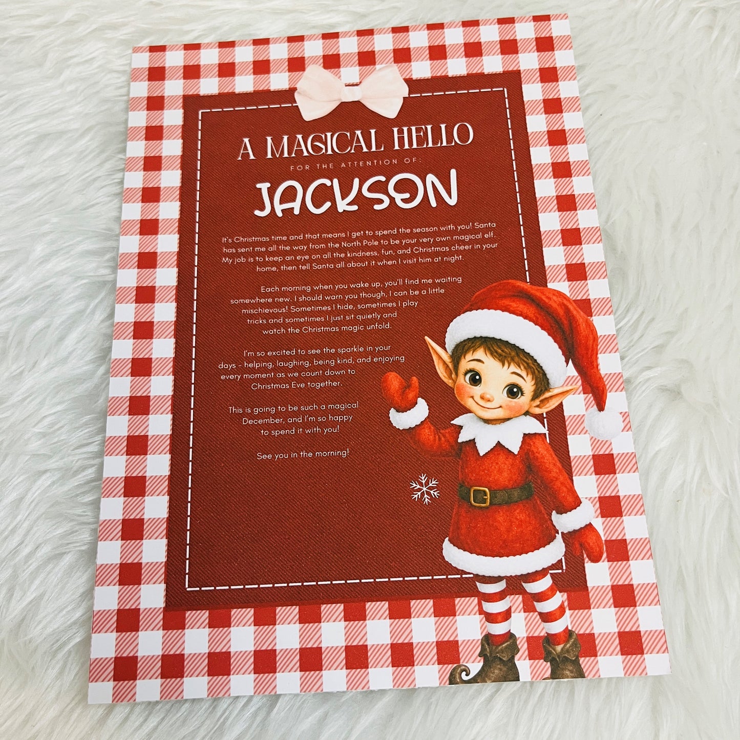 A personalised Christmas greeting card featuring a red and white gingham border, a dark red center panel with festive text, and an illustration of a elf character waving. The card is photographed flat on a white faux fur surface.