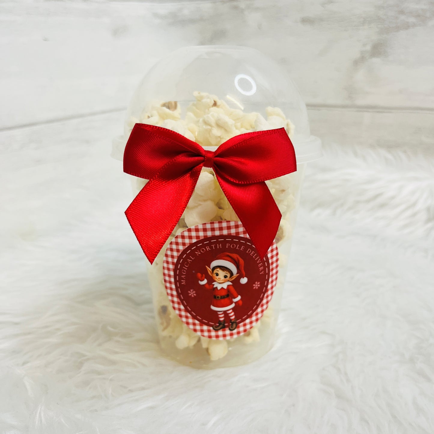 A clear plastic popcorn jar filled with white popcorn and tied with a bright red satin bow, featuring a round red-and-white holiday sticker with a cartoon elf and the text 'Magical North Pole Delivery'