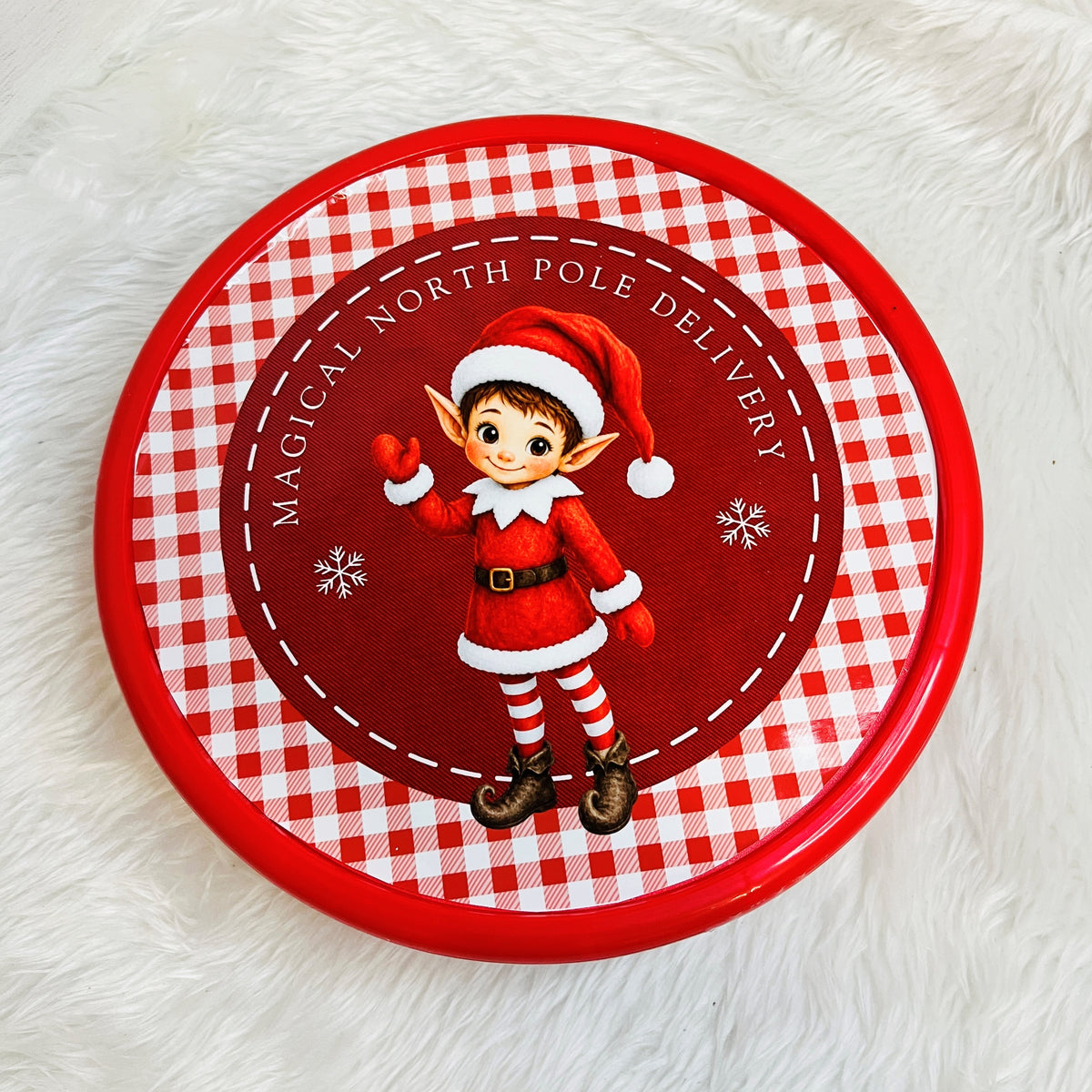 A round red and white gingham sweet tin featuring a cartoon elf in a Santa outfit with the text 'Magical North Pole Delivery' on a white faux fur background.
