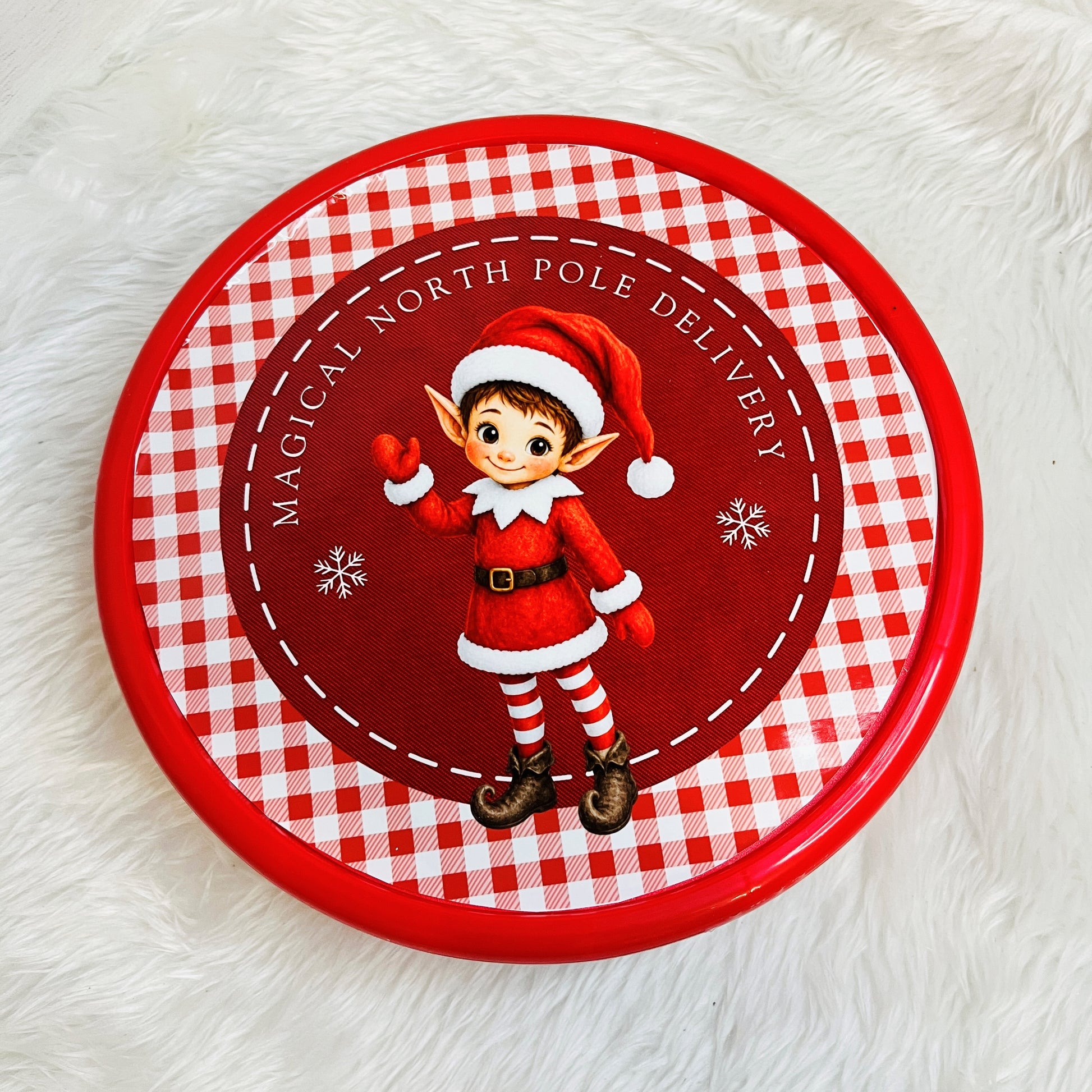 A round red and white gingham sweet tin featuring a cartoon elf in a Santa outfit with the text 'Magical North Pole Delivery' on a white faux fur background.