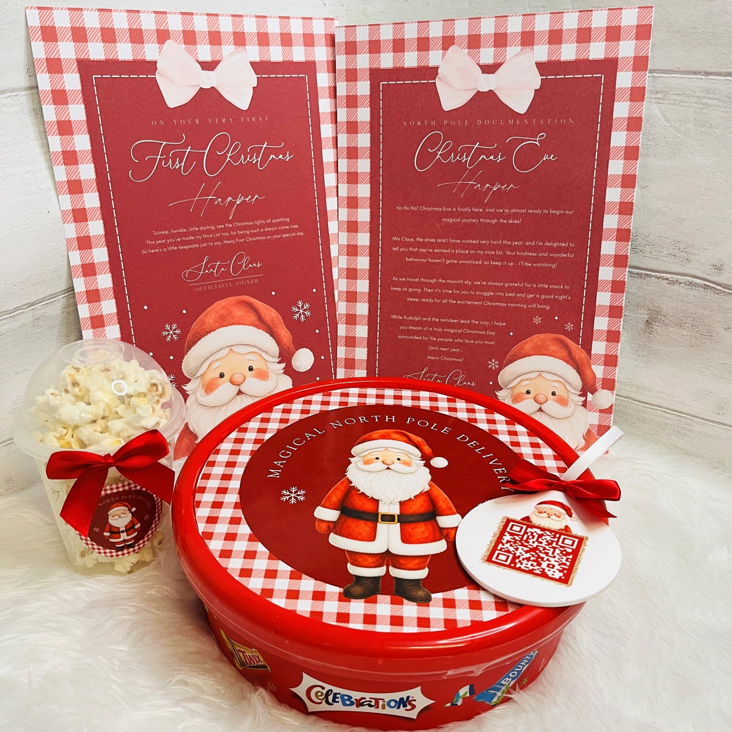 A Christmas-themed gift set featuring a round red tin with a Santa Claus illustration and a QR code tag, a clear popcorn bag tied with a red ribbon, and two red-and-white gingham personalised cards in the background.