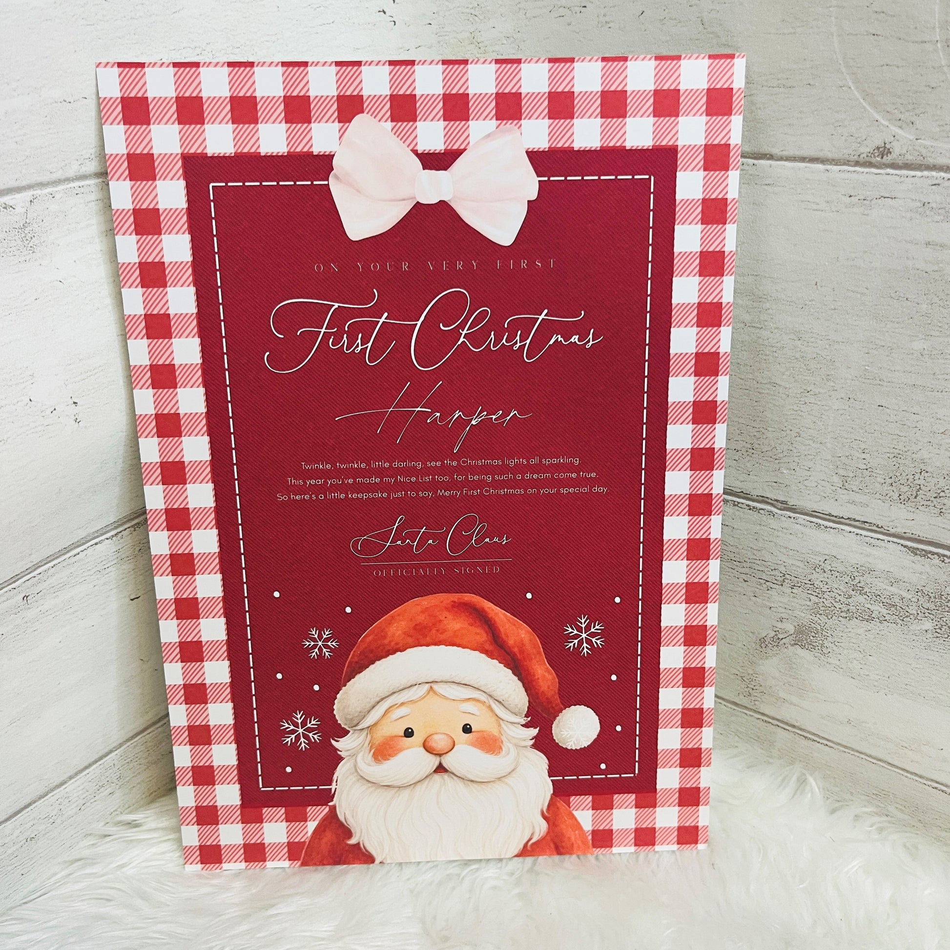 A personalised first Christmas card with a red and white gingham border, a pink bow illustration, and a Santa Claus illustration at the bottom, displayed on a white faux fur surface against a light gray wood background.