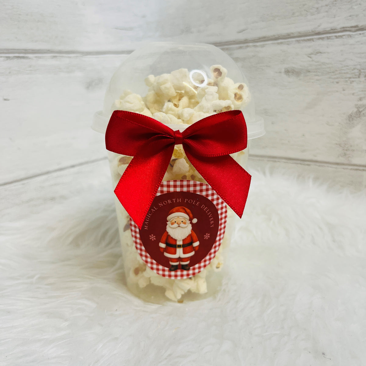 A clear plastic popcorn cup filled with white popcorn and tied with a large red satin bow, featuring a round Santa-themed label that reads 'Magical North Pole Delivery'. The cup is placed on a white faux fur surface against a light gray wood plank background.