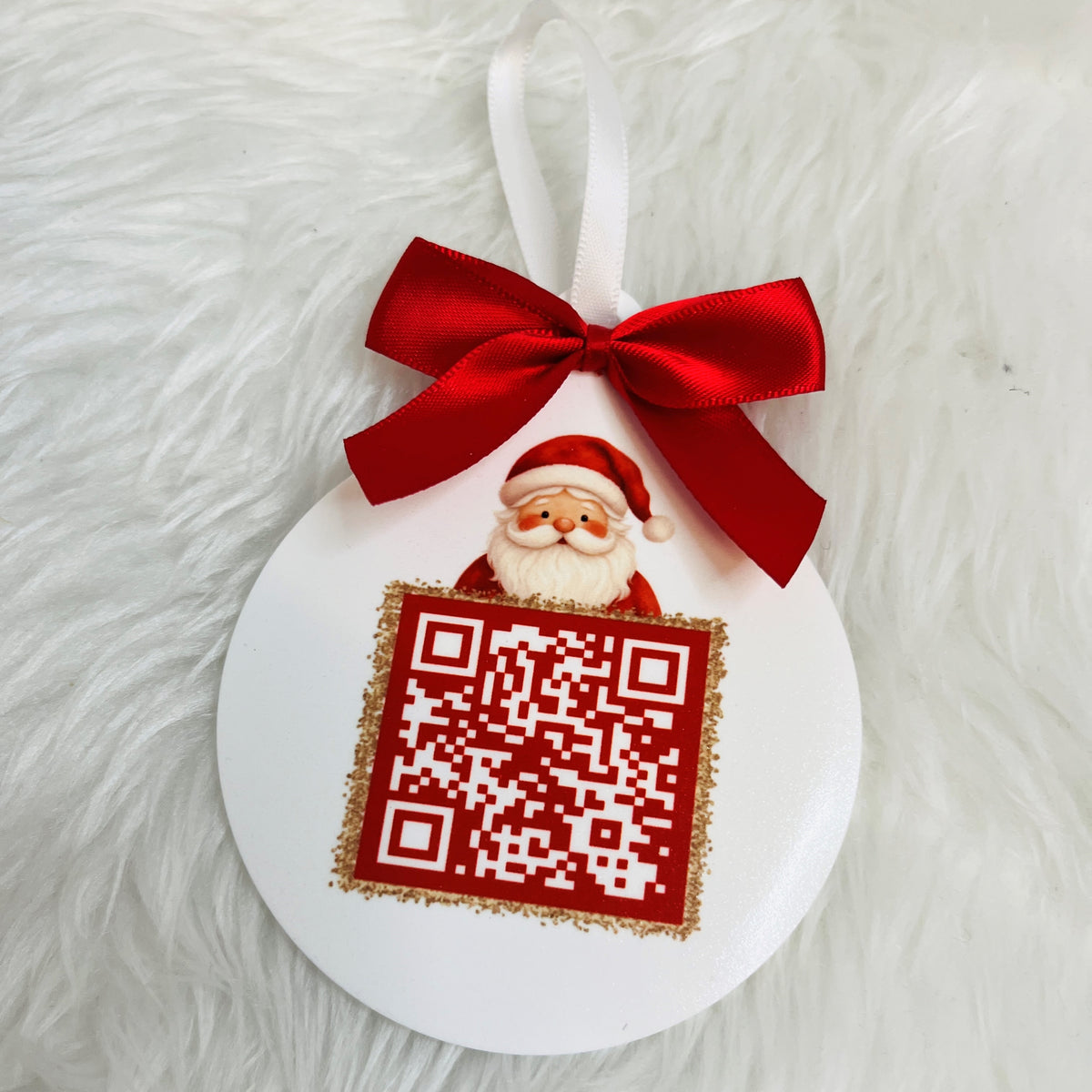 A white round Christmas ornament with a red ribbon loop, a red bow, a small Santa Claus figure, and a red square panel with a QR code, placed on a white faux-fur surface.