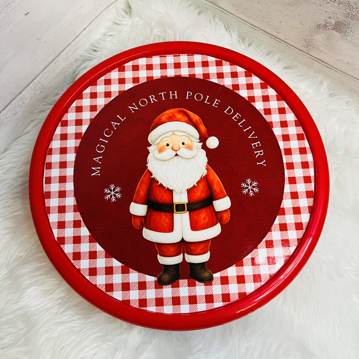 A round red and white gingham tin lid featuring a cartoon Santa Claus and the text 'MAGICAL NORTH POLE DELIVERY', placed on a white faux fur surface.