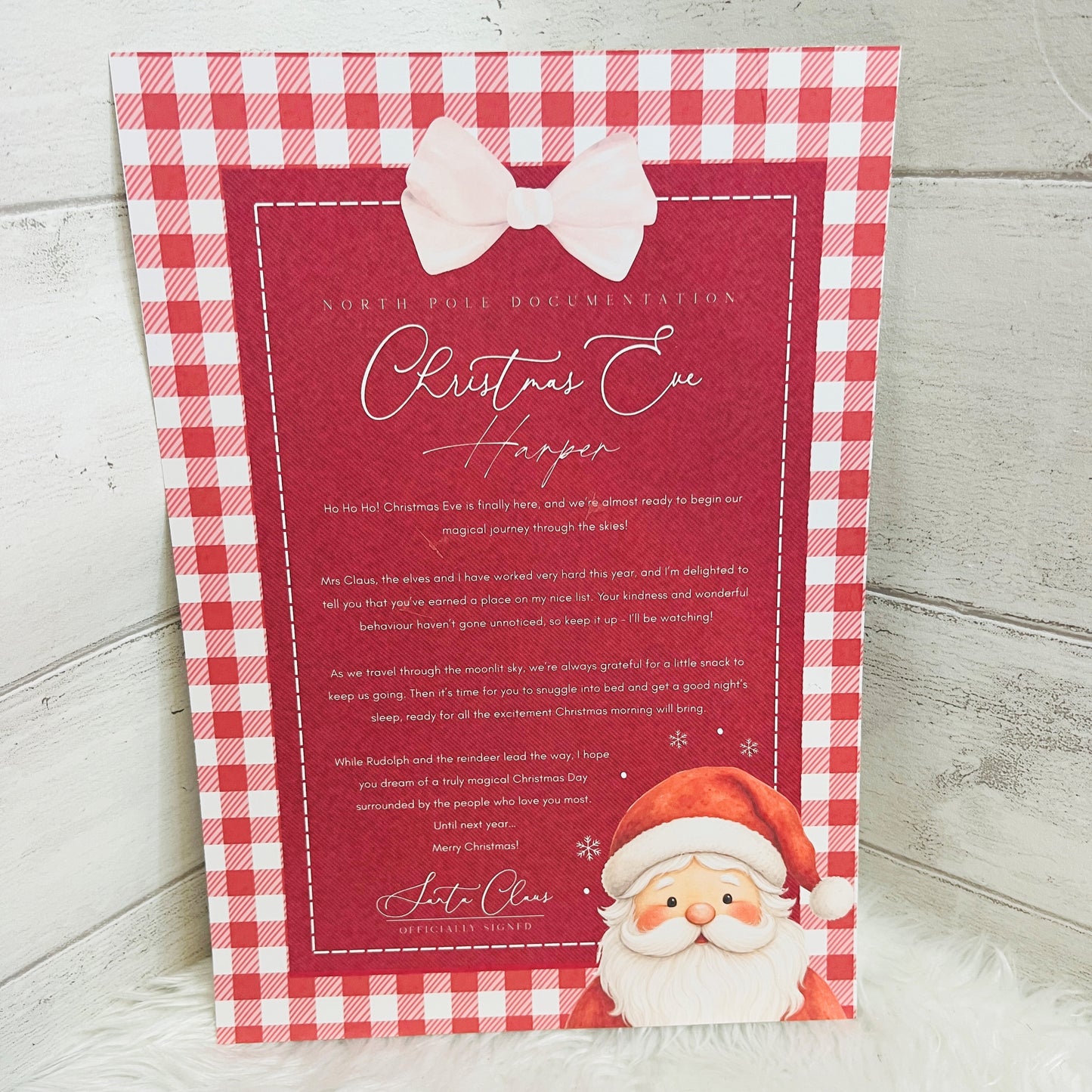 A personalised Christmas Eve letter print with a red and white gingham border, a pink bow illustration, white script text, and a Santa Claus illustration in the bottom right corner, displayed against a light gray wood plank background.