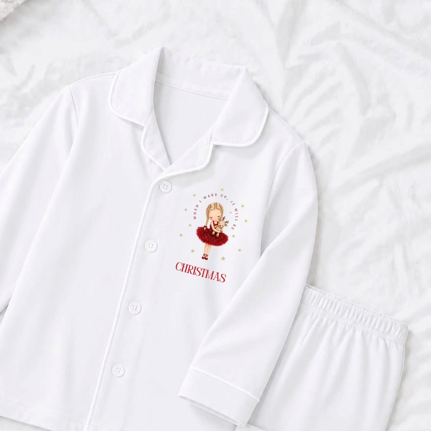 White long-sleeve pajama top and matching trousers laid flat on a white textured fabric background. The pajama top has a collar, front buttons, and a small Christmas-themed graphic on the left chest with the word 'When i wake up it will be christmas' below it.