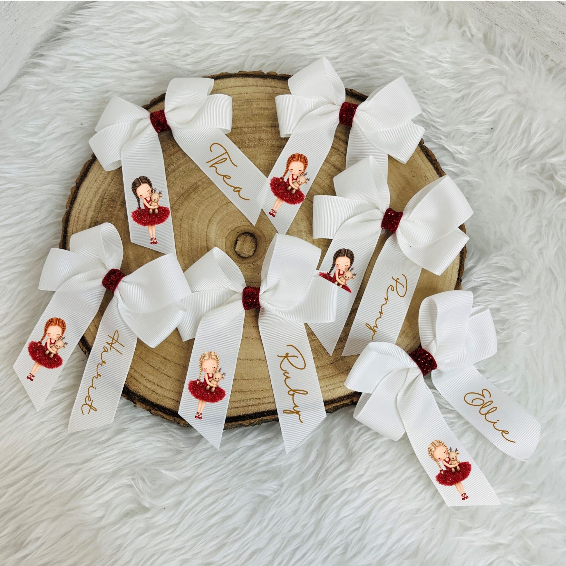 personalised white hair bows arranged on a round wood slice over a white faux fur surface. Each bow has a white ribbon tail printed with a custom portrait of a girl in a red dress, gold script names, and a small dark red glittery center.