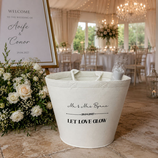 A off white felt bucket gift bag with custom wedding text and date, displayed on a marble floor in a reception venue next to a floral arrangement and a welcome sign.