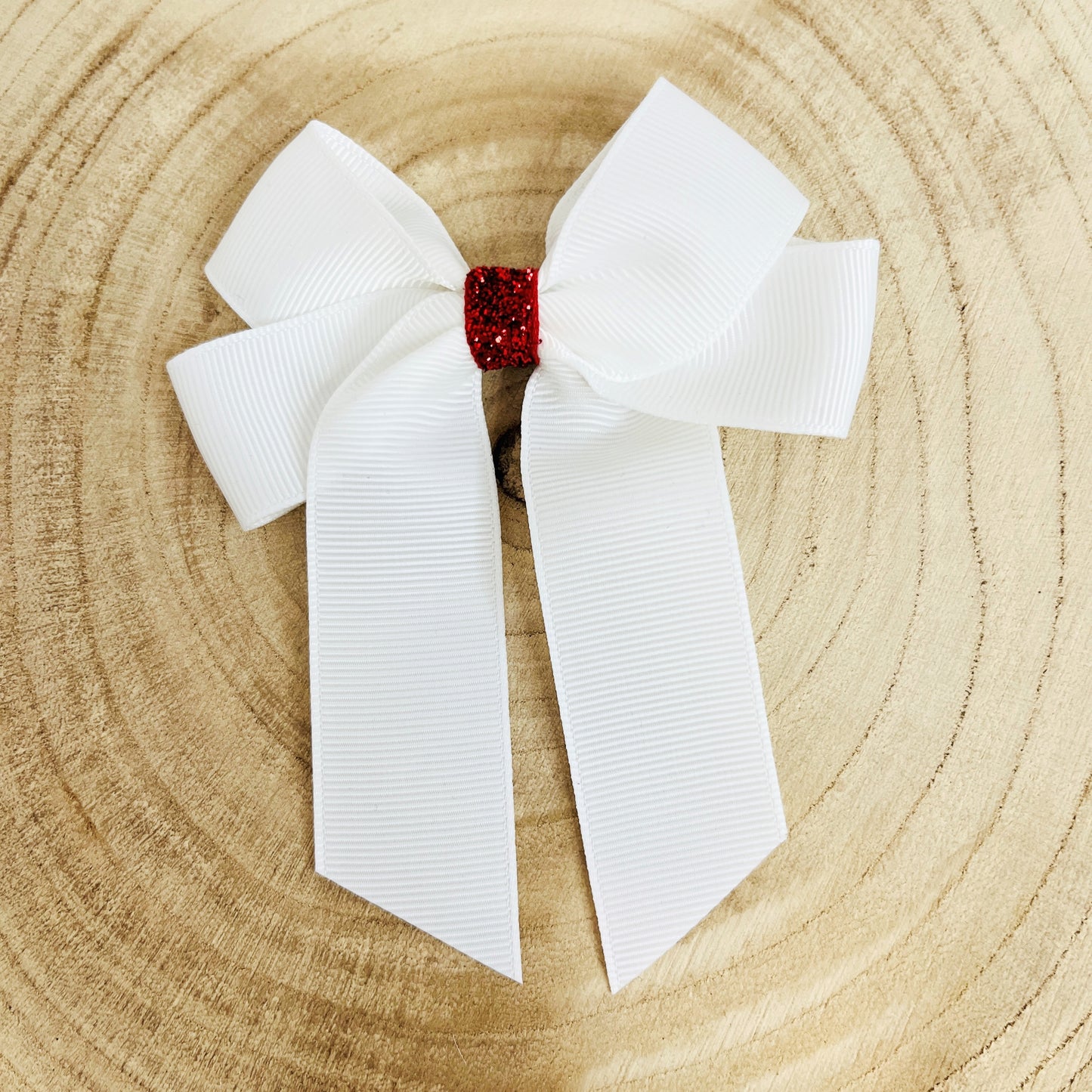 A white grosgrain ribbon hair bow with a small red glittery center, displayed flat on a light wood slice background.