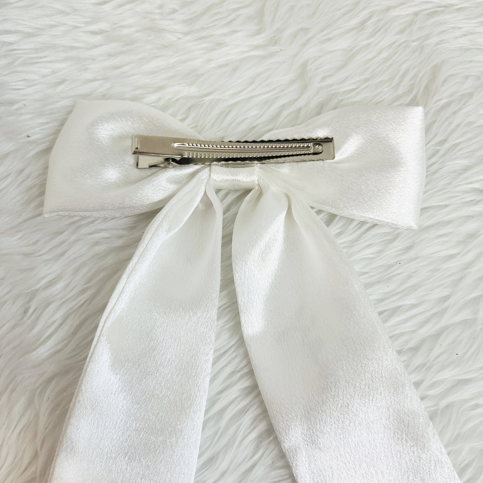A white satin bow hair clip with a silver metal clip, displayed on a white faux fur background.