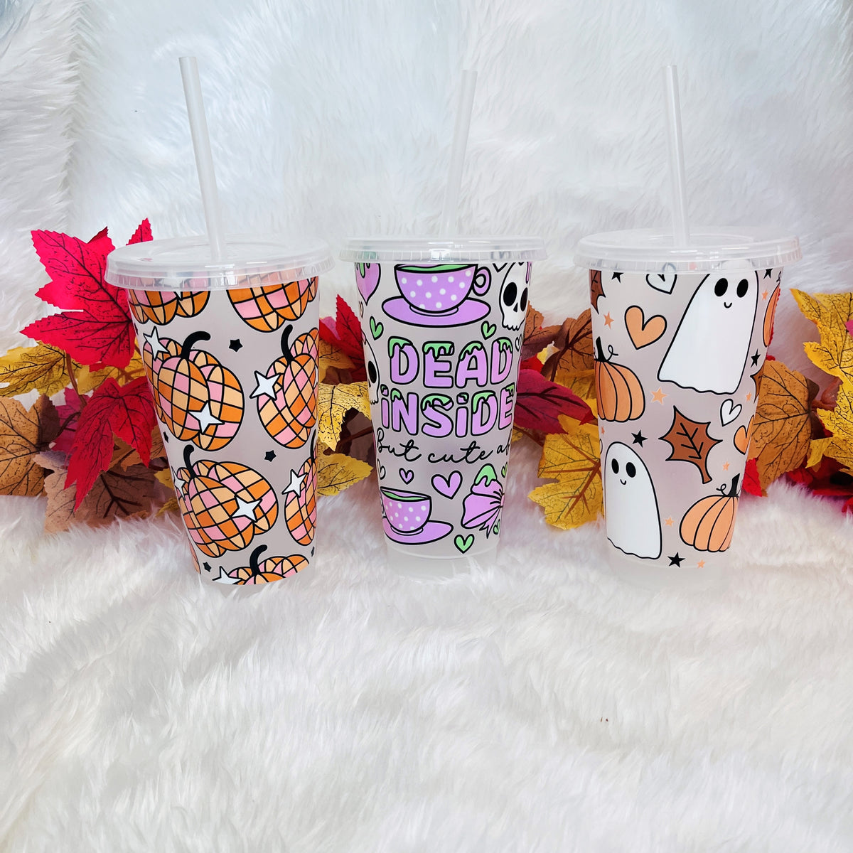 Three frosted plastic Halloween-themed tumblers with clear straws, decorated with pumpkins, ghosts and the phrase 'DEAD INSIDE', displayed on a white faux fur surface with autumn leaves.