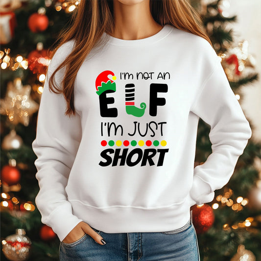 Woman wearing a white jumper with the graphic 'I'm not an elf, I'm just short' in front of a blurred christmas tree
