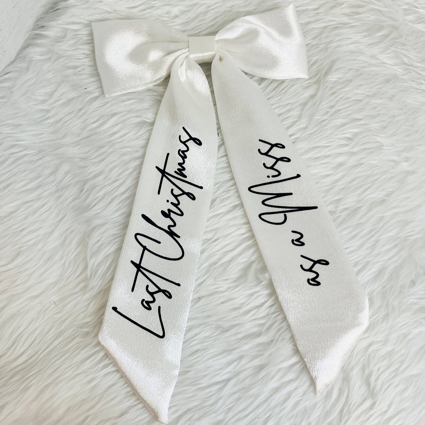 A white satin bow with long ribbon tails printed with black script text reading 'Last Christmas' on one side and 'Miss Whip' on the other, laid flat on a white faux fur background.