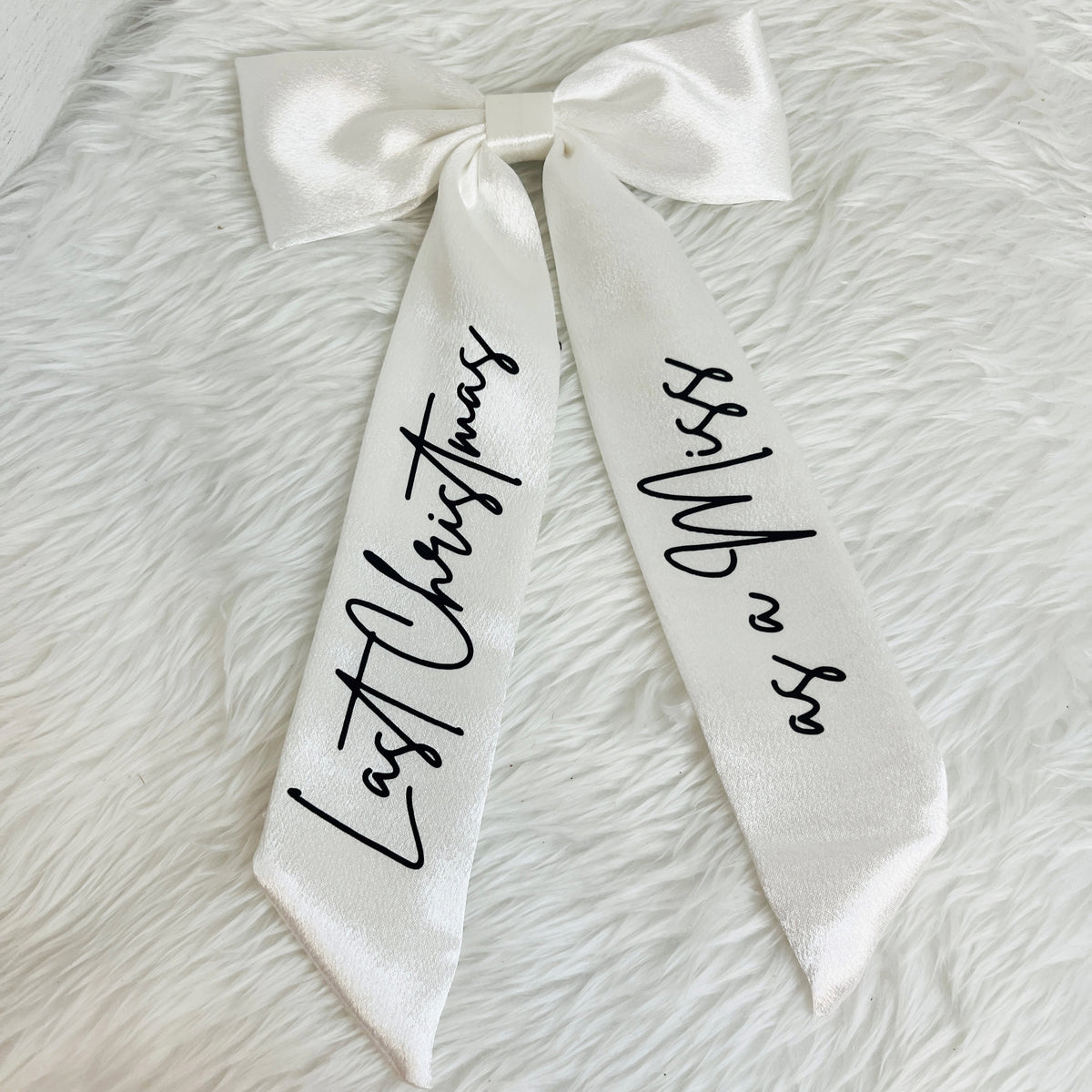 A white satin bow with long ribbon tails printed with black script text reading 'Last Christmas' on one side and 'Miss Whip' on the other, laid flat on a white faux fur background.