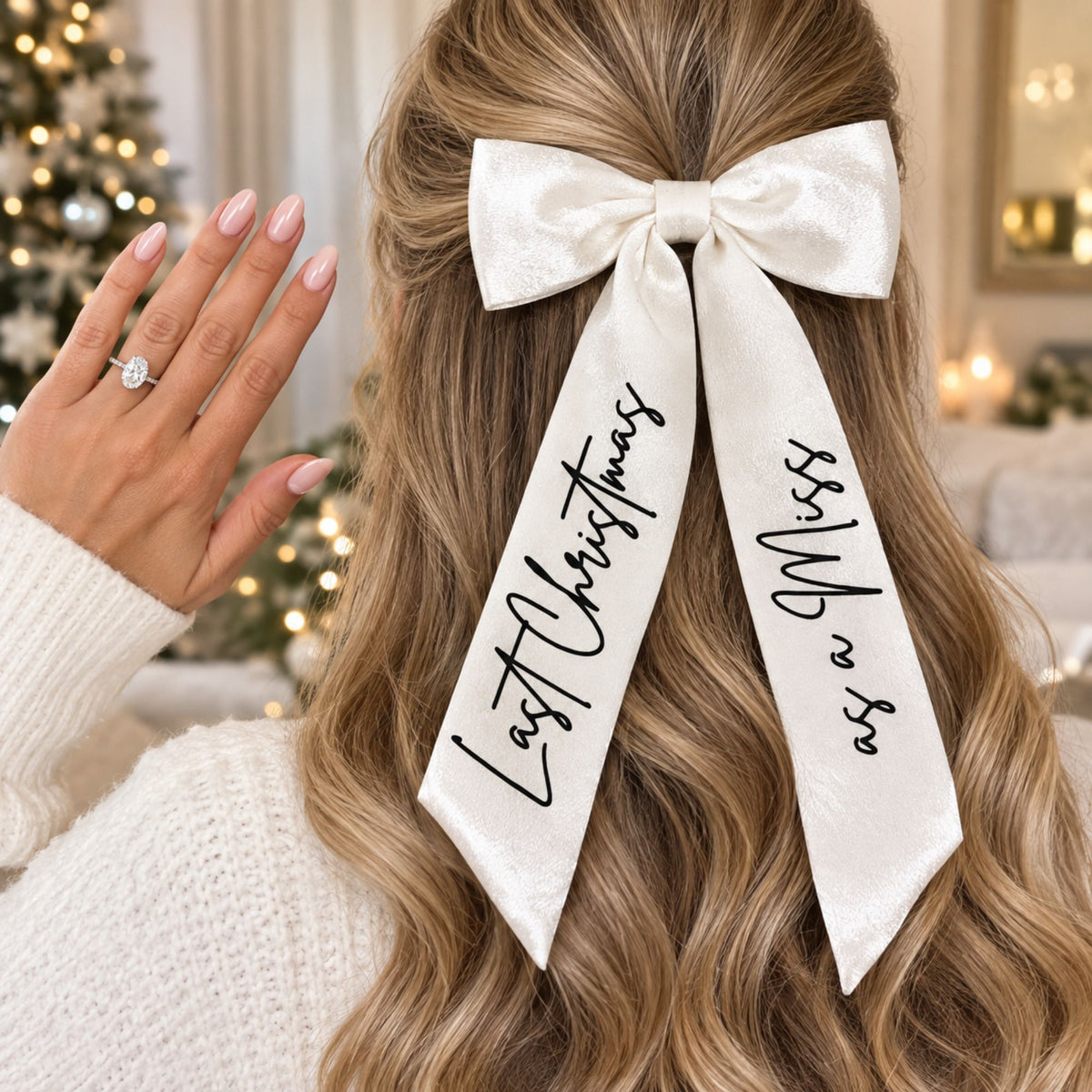 A woman with long wavy blonde hair wears a large white satin bow hair clip with black script text reading 'Last Christmas' and 'as a Miss'  in a cozy indoor christmas setting.