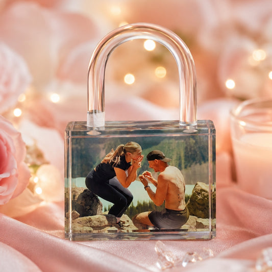 A clear crystal love lock with a photo with a printed photo of a couple in a mountain setting, displayed on pink satin fabric with blurred pink roses and warm lights in the background.