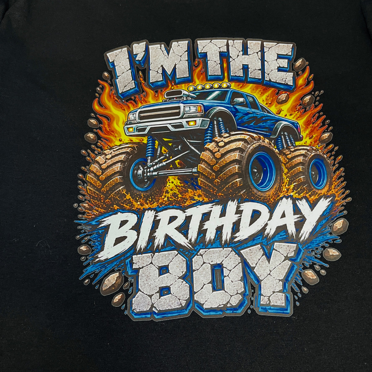 A close-up of a black t-shirt featuring a large graphic print that reads 'I'M THE BIRTHDAY BOY' in white and blue lettering, surrounding an illustration of a blue monster truck with large off-road tires and flames.