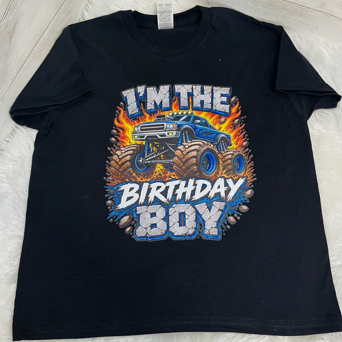A black short-sleeve t-shirt laid flat on a white faux fur surface, featuring a colorful graphic of a blue monster truck with flames and the text 'I'M THE BIRTHDAY BOY'.