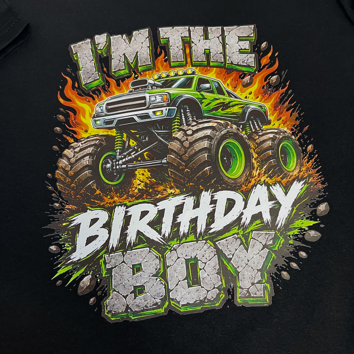 A close-up of a black t-shirt featuring a vibrant graphic print. The design shows a green and black monster truck surrounded by orange flames, with the text 'I'M THE BIRTHDAY BOY' in a bold, cracked stone-style font with green outlines.