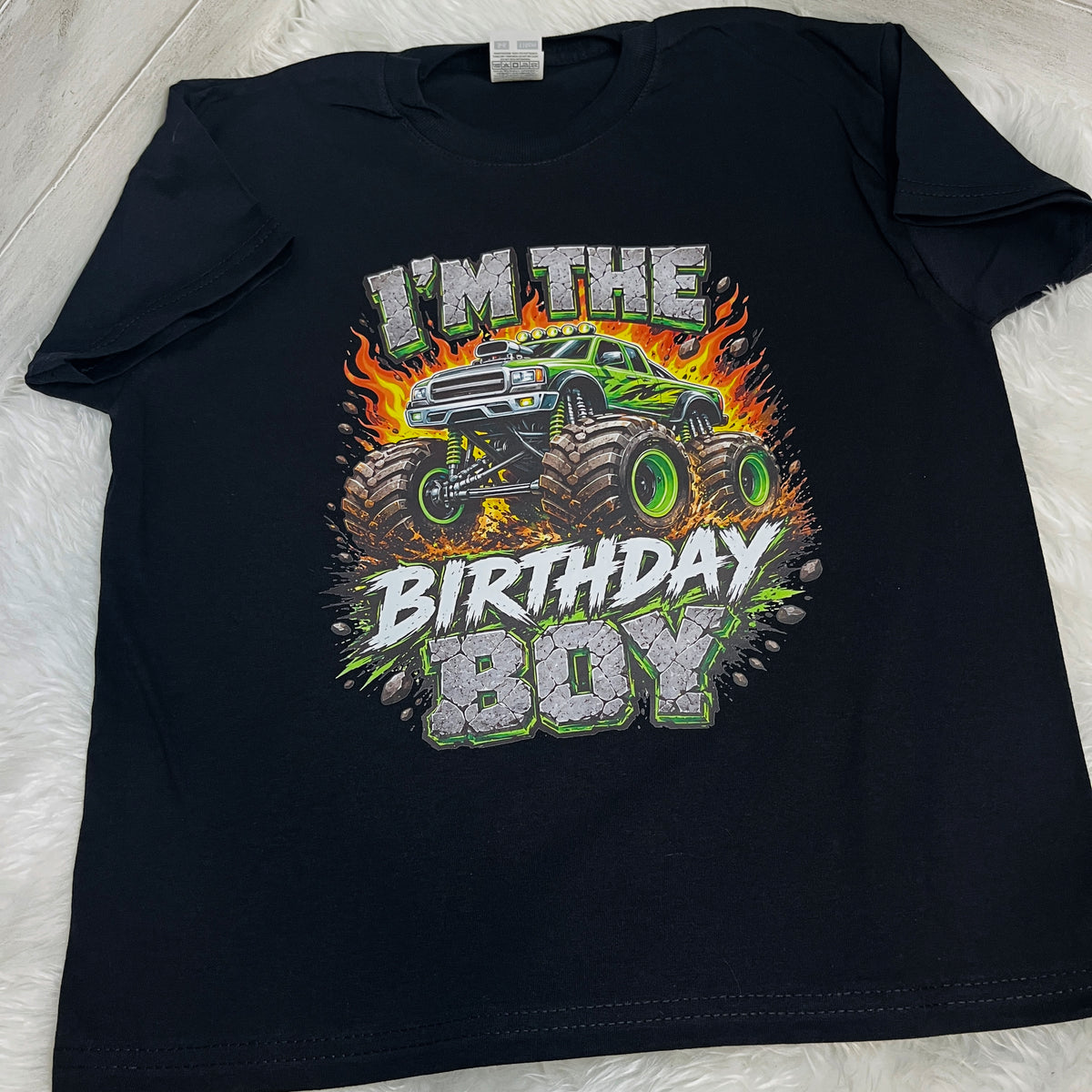 A black short-sleeve t-shirt laid flat on a white textured surface, featuring a colorful graphic of a green monster truck with flames and the text 'I'M THE BIRTHDAY BOY'.