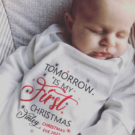 A sleeping newborn baby wearing a white long-sleeve sleepsuit with a personalised Christmas graphic that reads 'TOMORROW IS MY First CHRISTMAS' in black and red script with star accents.