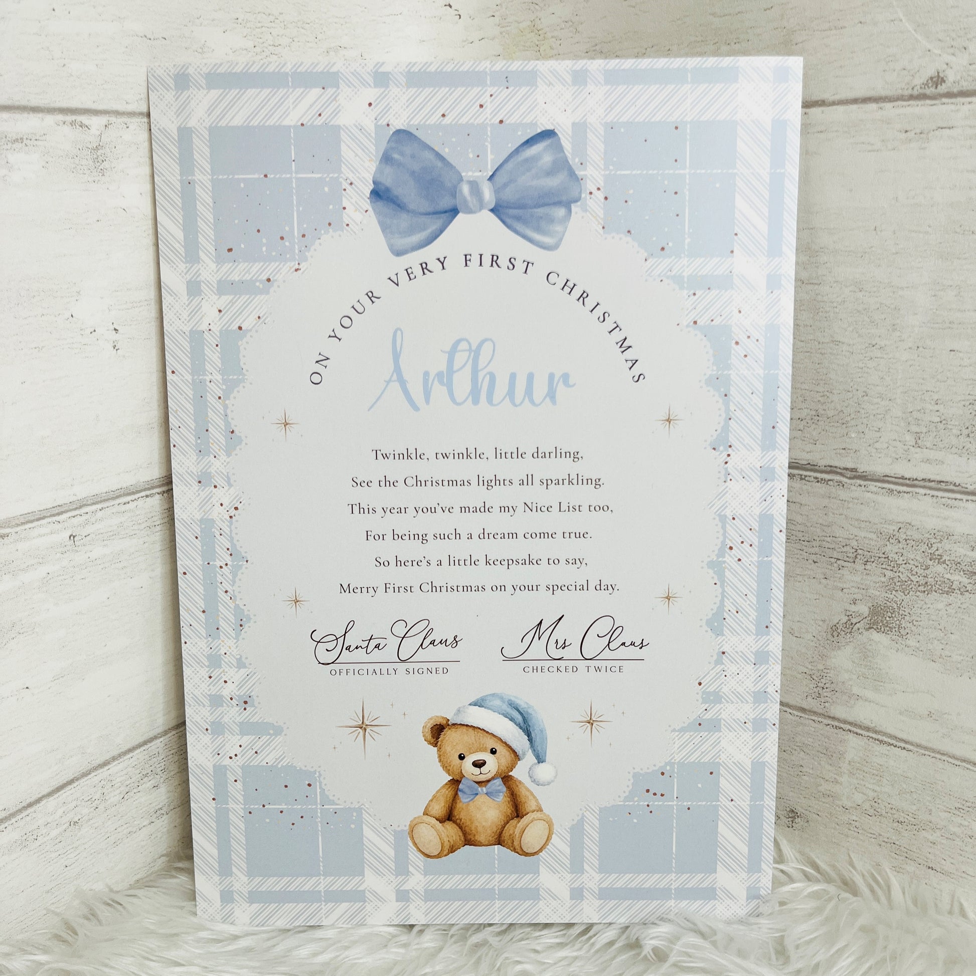A personalised A4 Christmas greeting card featuring a light blue plaid border, a blue bow illustration, a teddy bear in a Santa hat, and a custom message for a child named Arthur, displayed on a white textured surface against a light wood background.