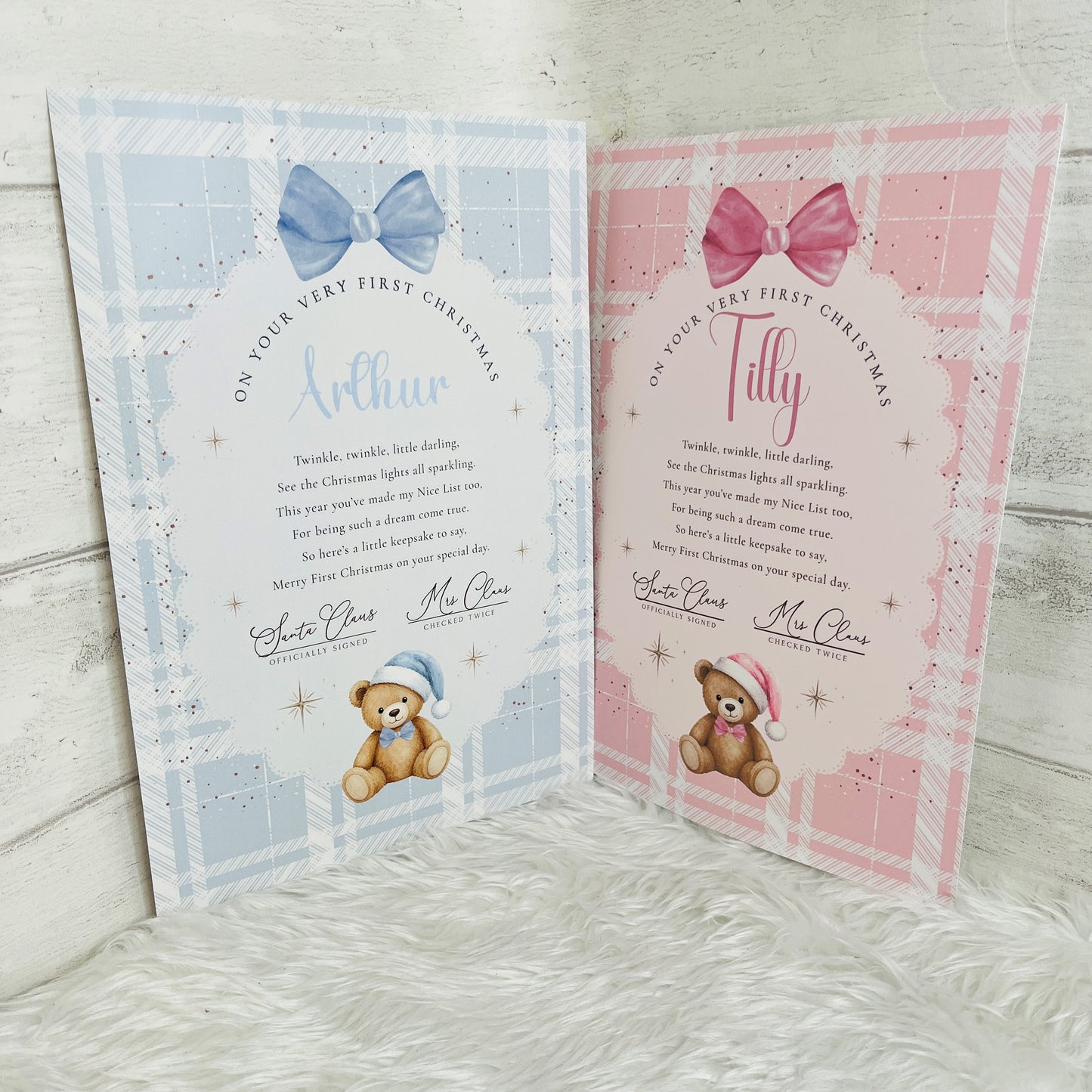 Two personalised baby first Christmas cards displayed side-by-side on a white faux fur surface against a light gray wood background. The left card is light blue with a blue bow and a teddy bear wearing a blue hat, personalized with the name 'Arthur' and the pink card is personalised with the name 'Tilly' in pink