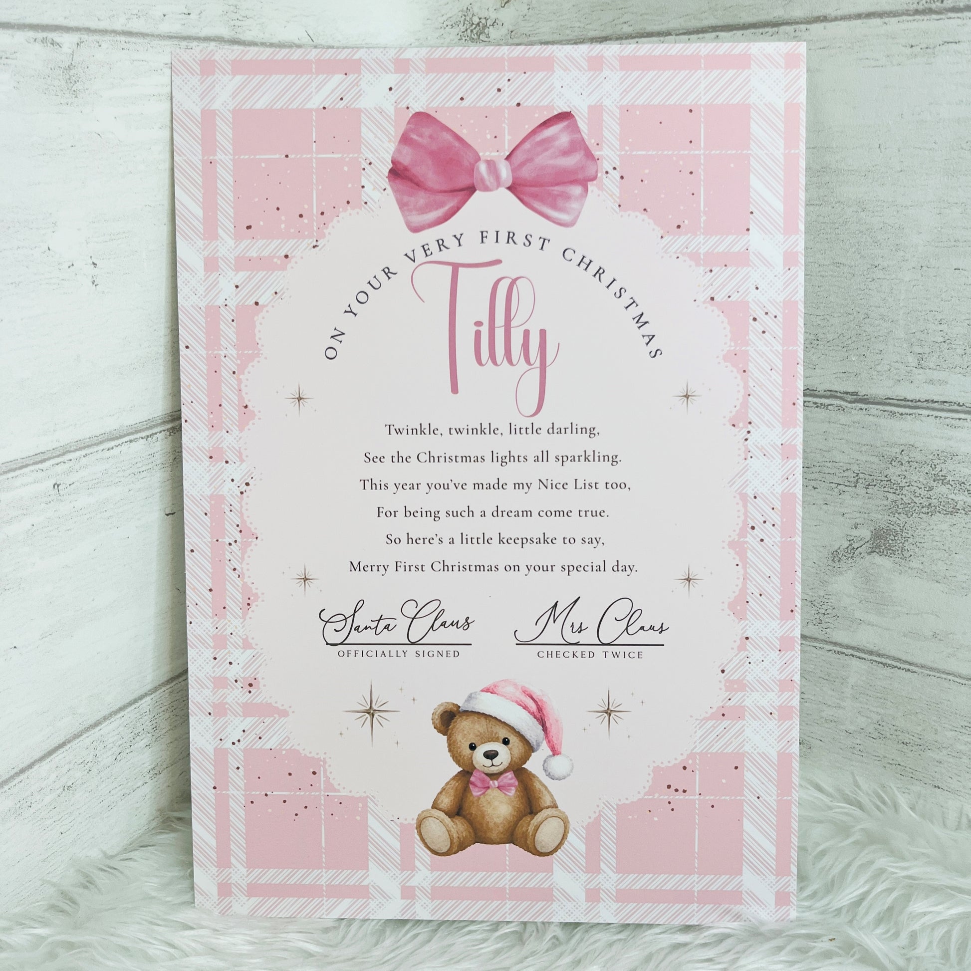 A personalised A4 Christmas card with a pink plaid border, a pink bow at the top, and a teddy bear wearing a Santa hat at the bottom. The card reads 'On your very first Christmas' and is signed by Santa Claus and Mrs Claus