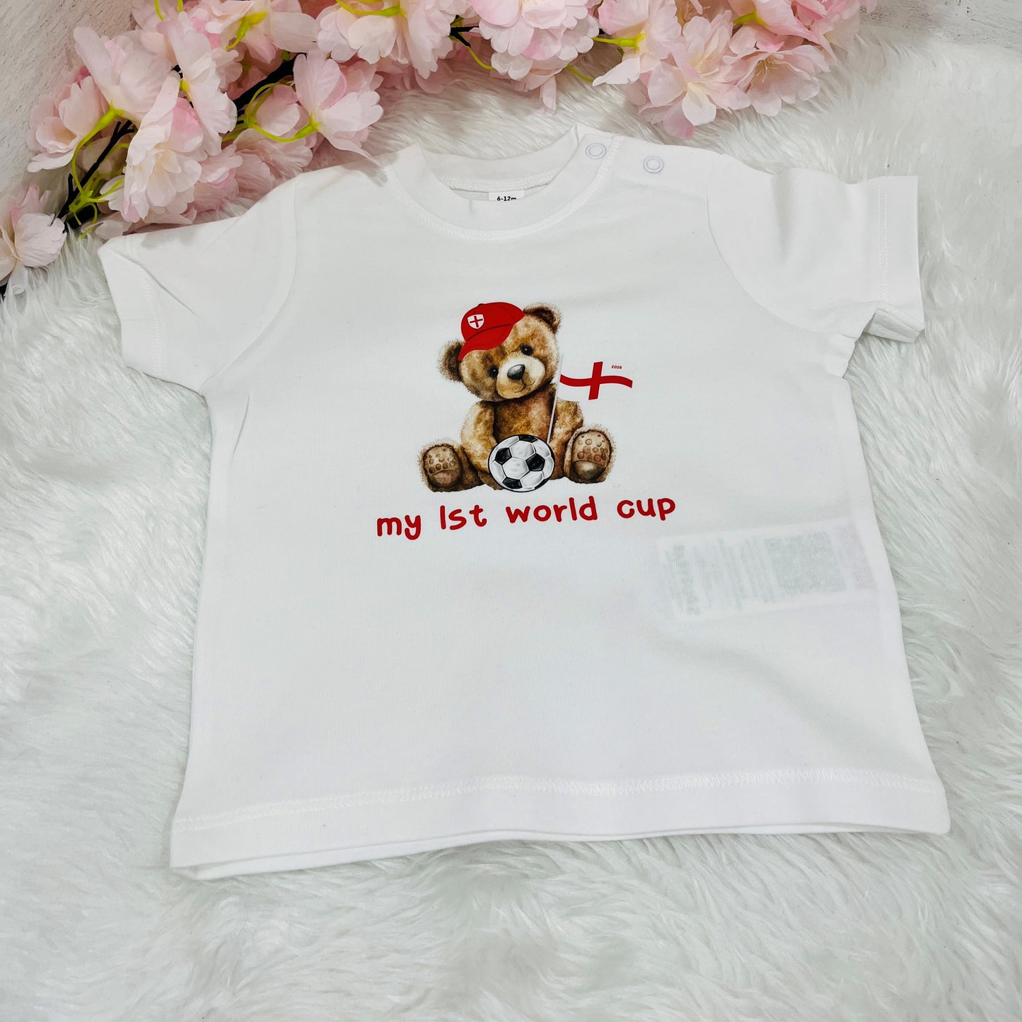 White t-shirt with a teddy bear design and 'My First World Cup' text on a soft surface with flowers in the background