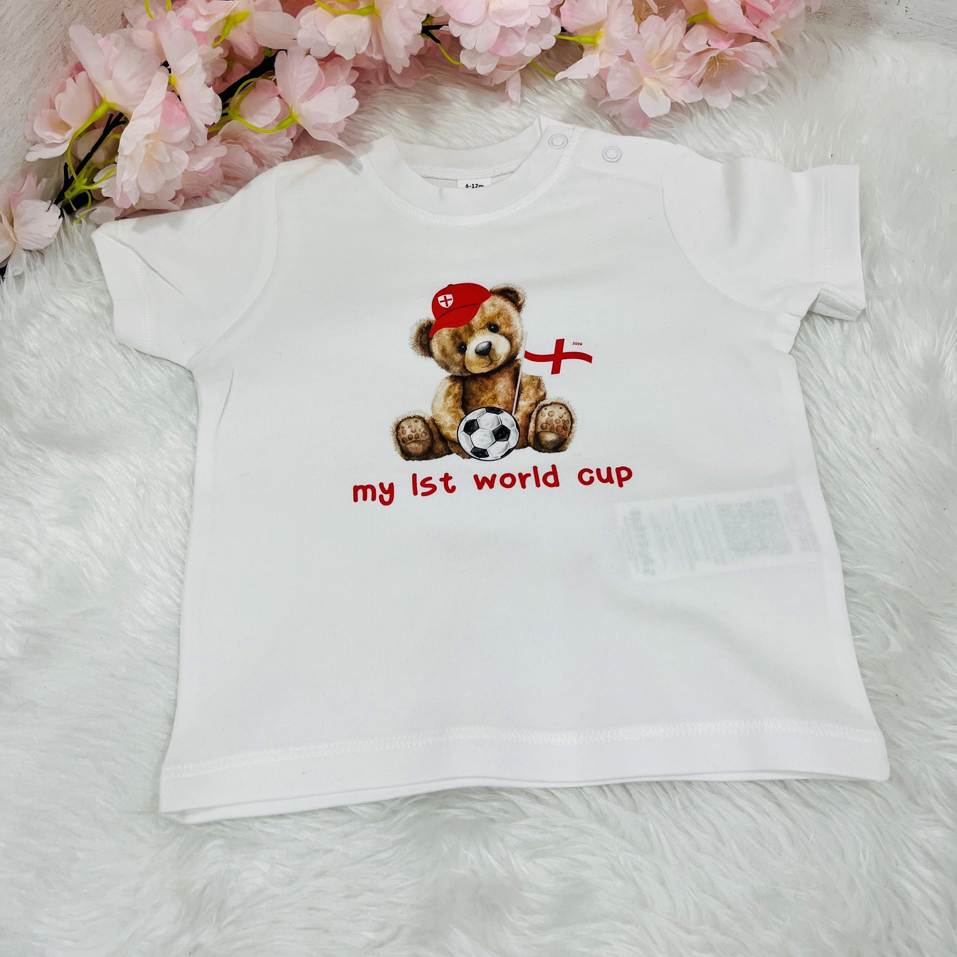 White t-shirt with a teddy bear design and 'My First World Cup' text on a soft surface with flowers in the background