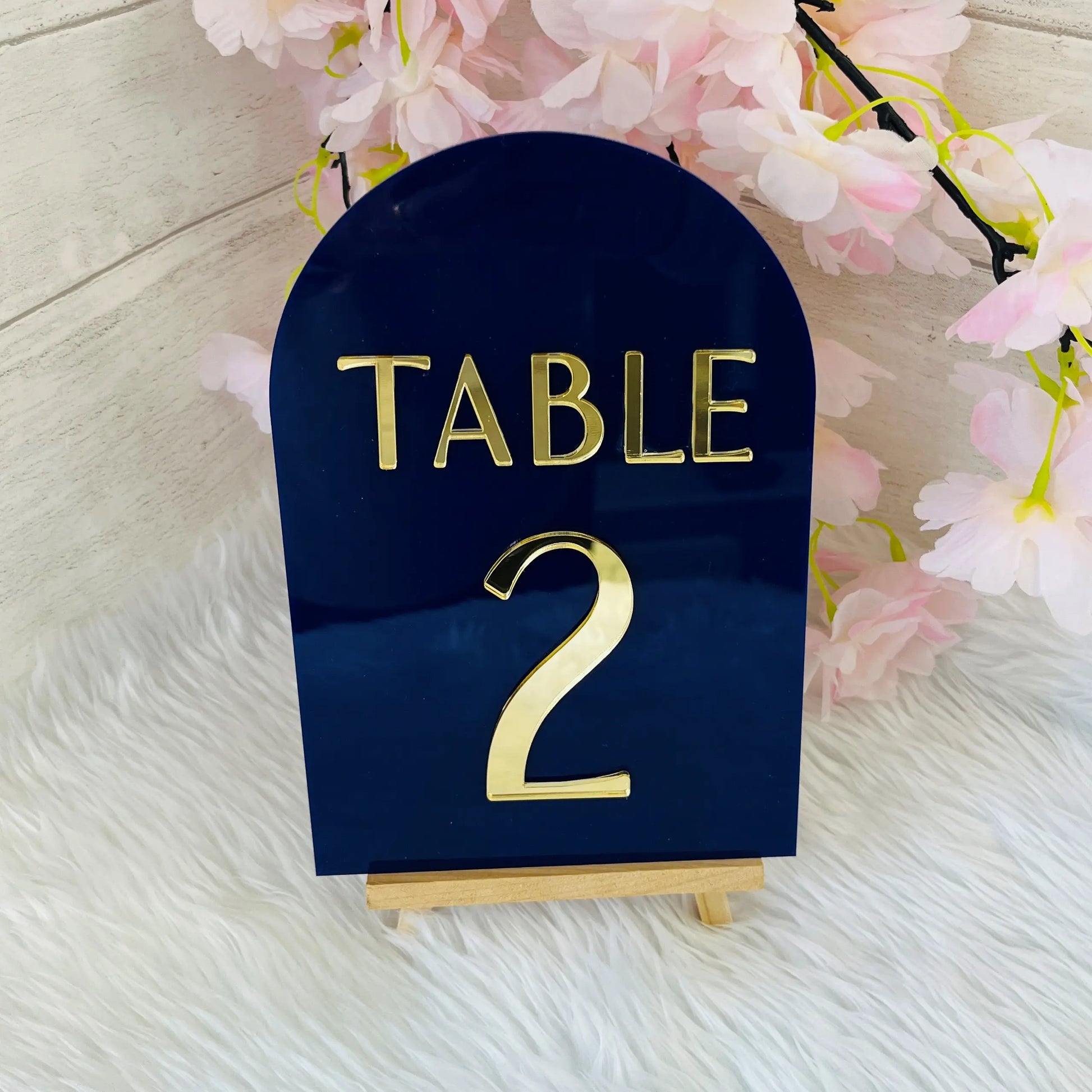 Navy blue table number holder with gold 'TABLE 2' on a light wooden surface with pink flowers.