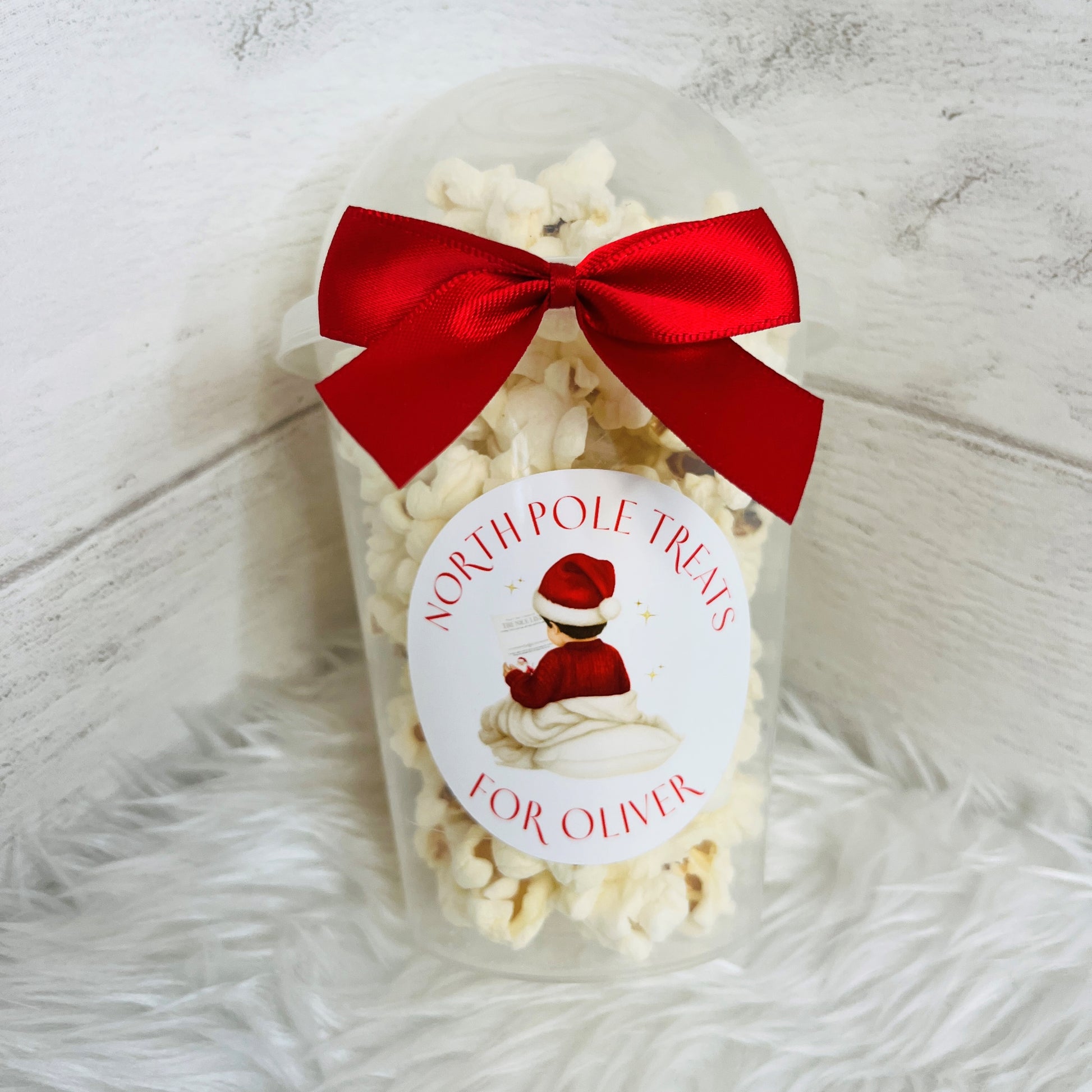 one clear plastic popcorn cups filled with white popcorn, each tied with a large red satin bow and a round white label featuring a child in a red jumper. The labels read 'NORTH POLE TREATS' and 'FOR OLIVIER' 