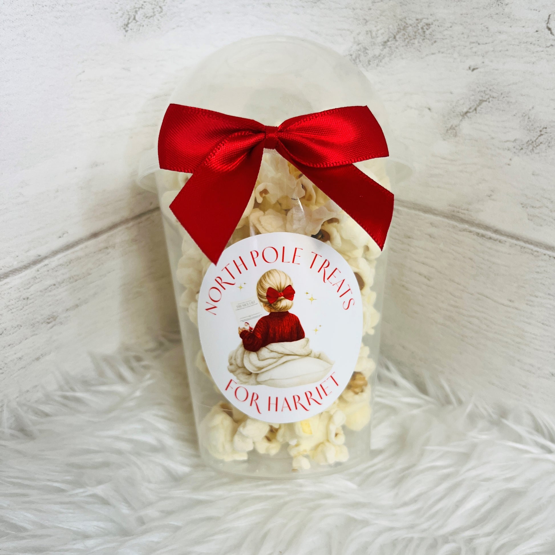 One clear plastic popcorn cups filled with white popcorn, each tied with a large red satin bow and a round white label featuring a young girl in a red jumper. The labels read 'NORTH POLE TREATS' and 'FOR HARRIET'.