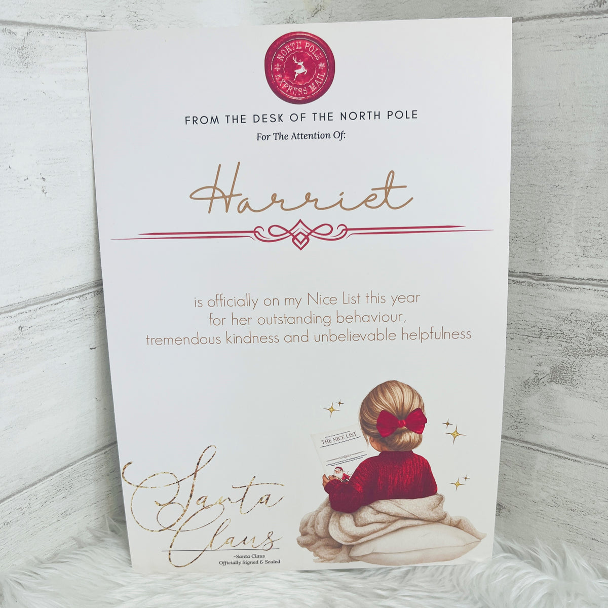 An A4 printed Nice list certificate addressed to 'Harriet' with a red North Pole seal at the top, gold script lettering, and an illustration of a little girl in a red jumper with a red bow in her hair sitting on a cushion holding a Nice List Certificate. The card is photographed leaning against a light wood surface on a white faux-fur surface.