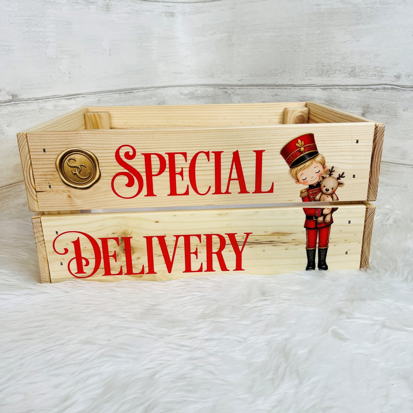 A light natural wood crate with 'SPECIAL DELIVERY' printed in red script on the front, featuring a nutcracker illustration and a gold seal emblem, set on a white faux fur surface against a white wood plank background.