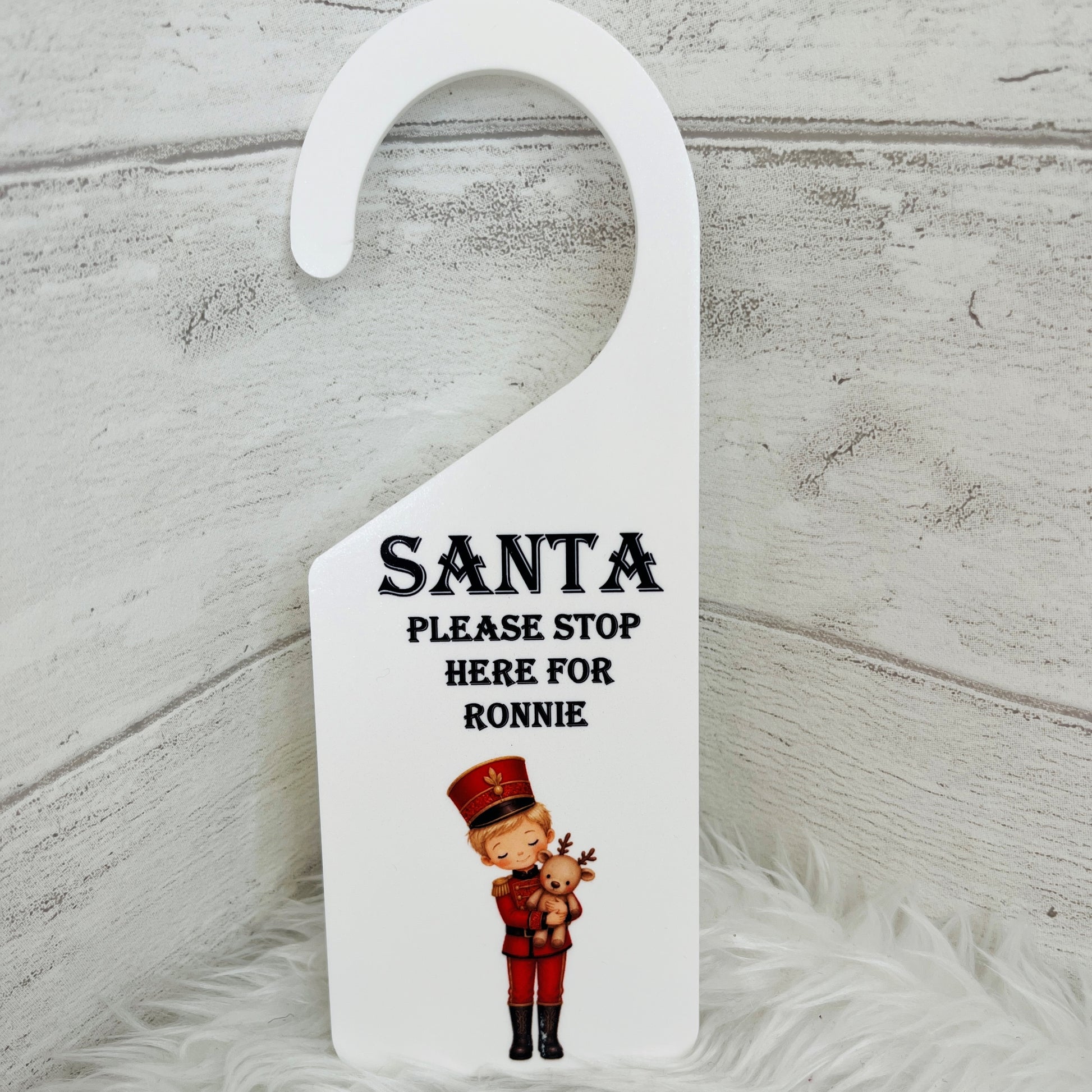 A white Christmas door hanger with the text 'SANTA PLEASE STOP HERE FOR RONNIE' and an illustration of a nutcracker holding a reindeer, set against a rustic white wood background.