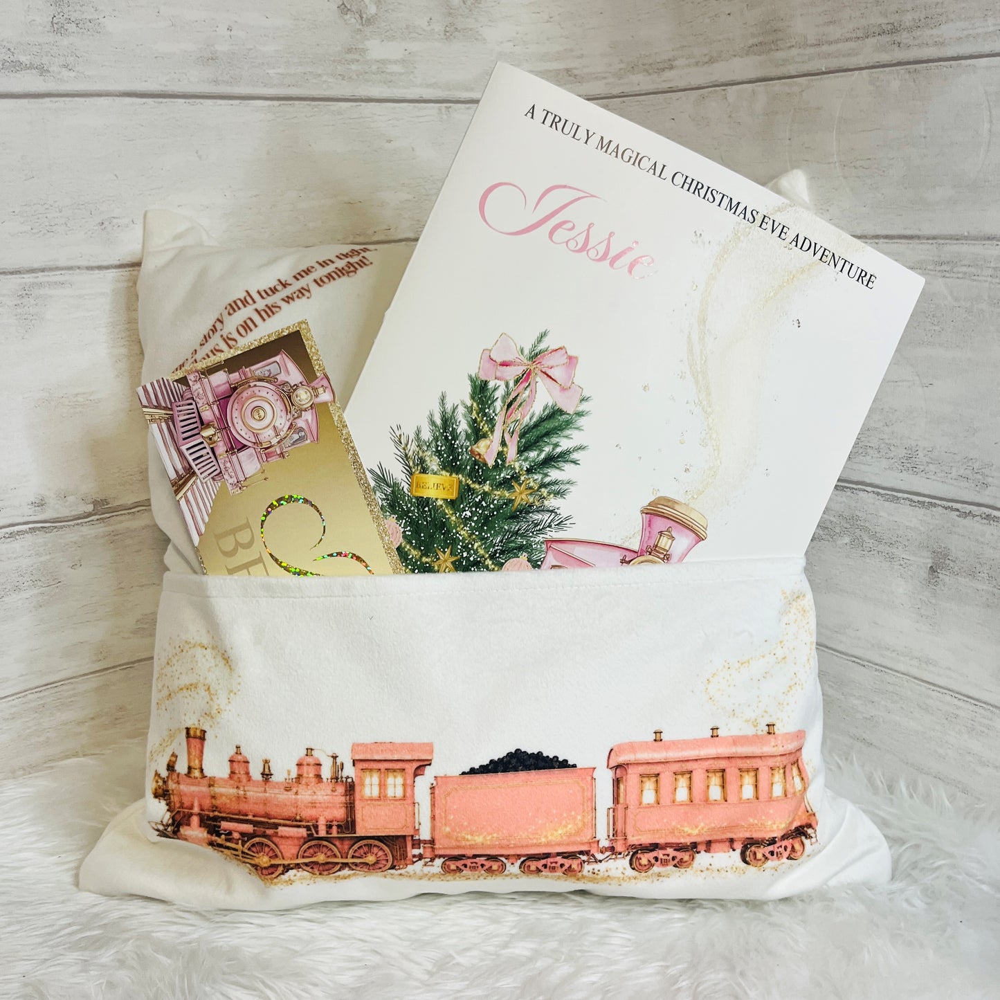A pink and gold Christmas gift pillow featuring a vintage-style pink train illustration on the front, styled with a personalised Christmas story book  and a small gold ticket on a white faux fur surface against a light wood plank background.