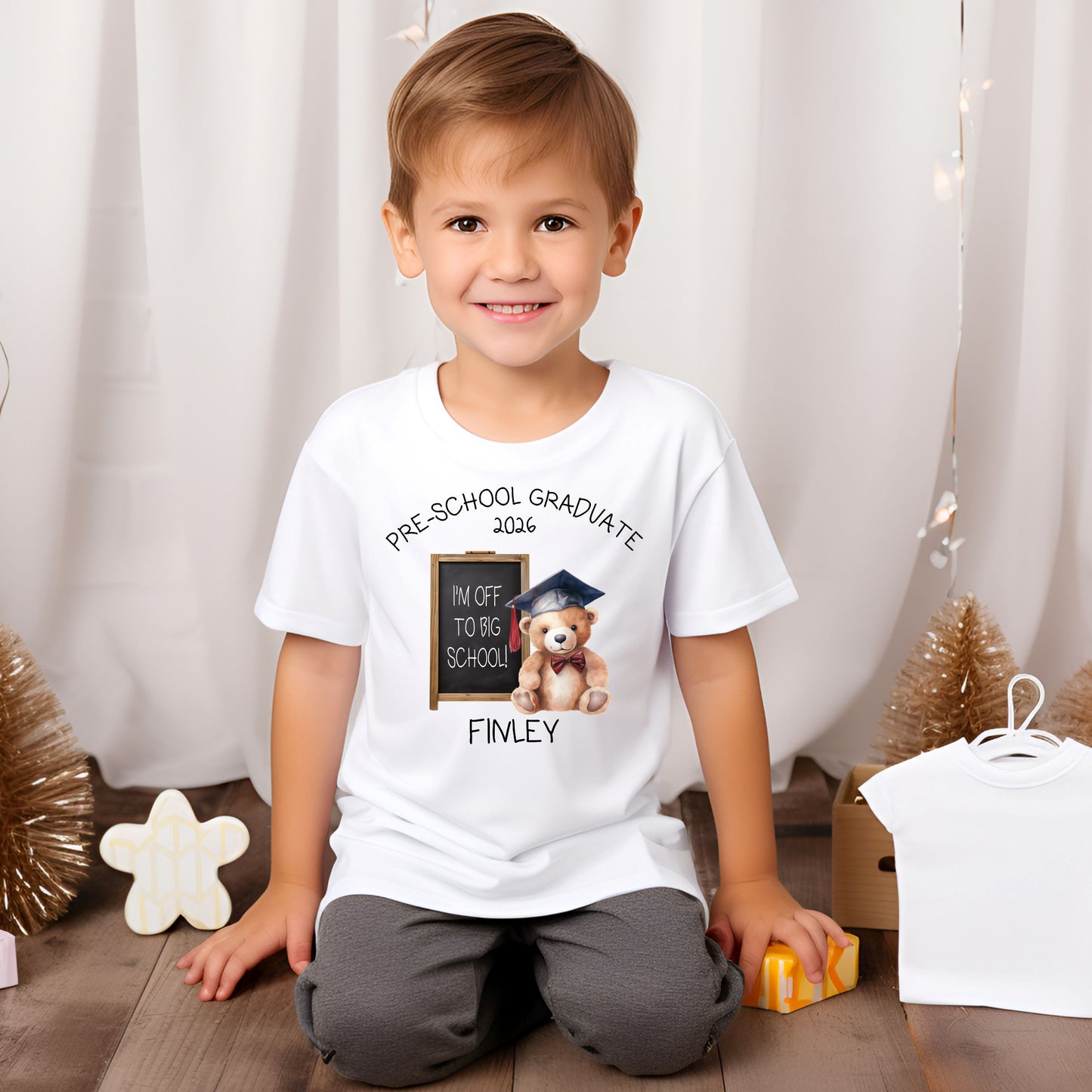 Child wearing a white t-shirt with a graduation design and text, sitting on the floor with a white curtain background.