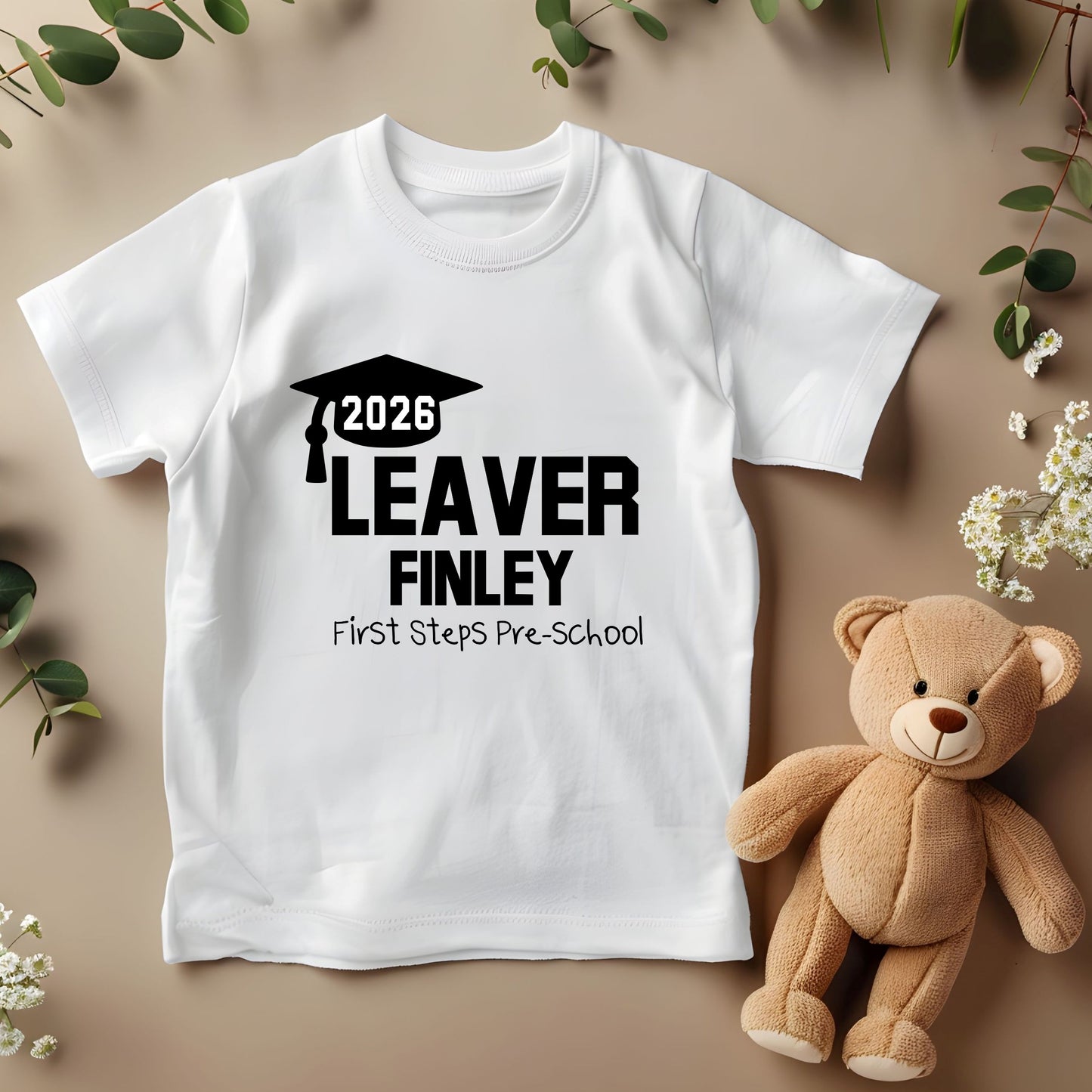 White t-shirt with '2026 Leaver Finley First Steps Pre-School' text, teddy bear, and flowers on a beige background