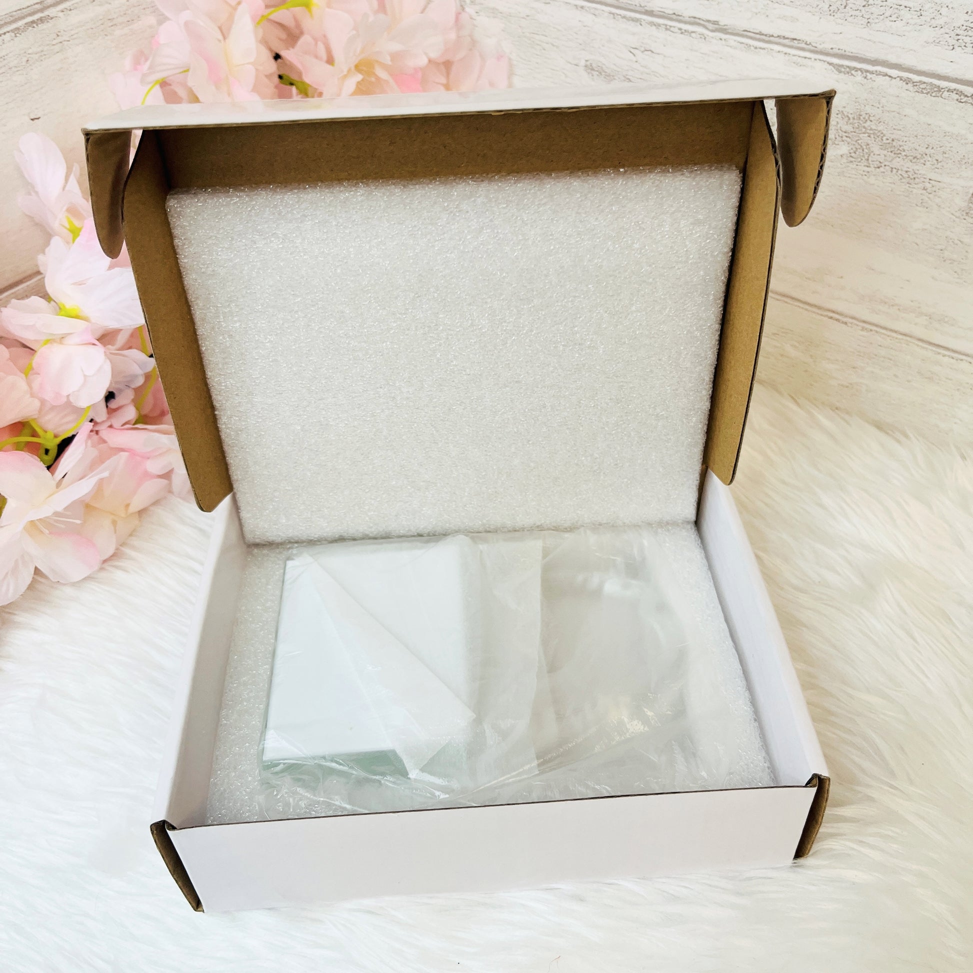 An open white cardboard box with a white foam insert, containing a crystal love lock, placed on a white furry surface with pink flowers in the background.