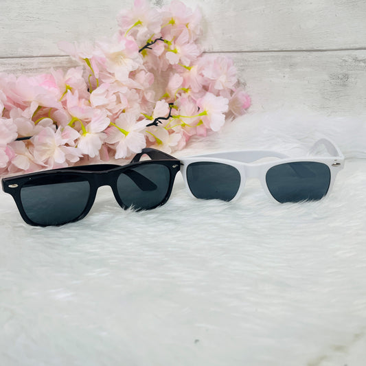 Two pairs of sunglasses, one black and one white, on a light wooden surface with pink flowers in the background.