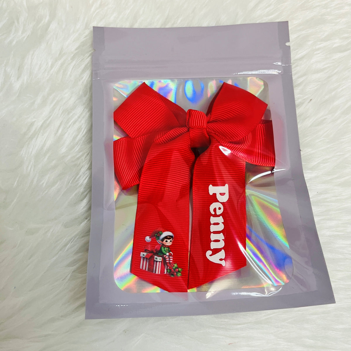 A red grosgrain ribbon bow with a vertical white printed name 'Penny' and a small Christmas elf graphic, presented inside a clear holographic zip pouch on a white faux fur background.