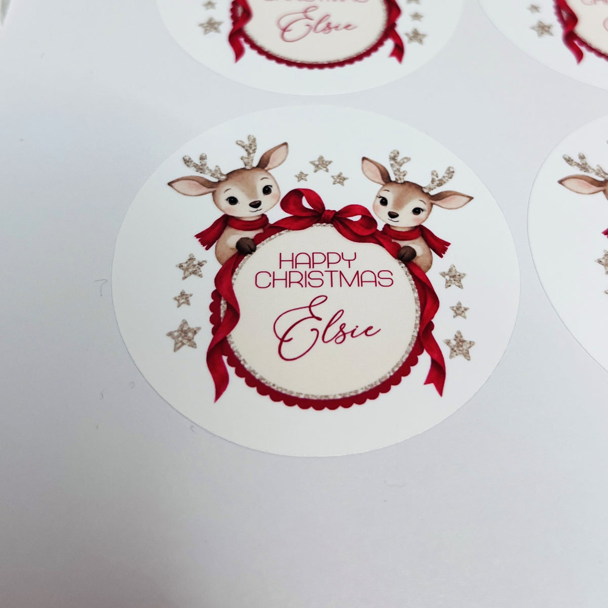 Round white Christmas sticker featuring two cartoon reindeer with red scarves, a large red bow, and the text 'Happy Christmas Elsie' in red script, shown on a white background with other partial stickers around the edges.
