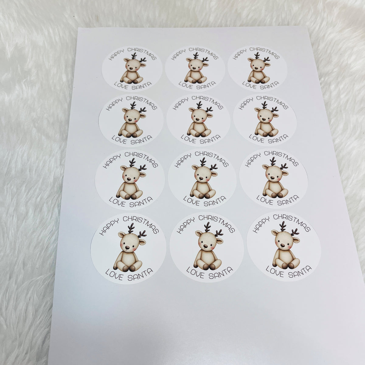 A sheet of round Christmas labels featuring a cute cartoon reindeer and the text 'HAPPY CHRISTMAS LOVE SANTA', displayed on a white faux fur surface.