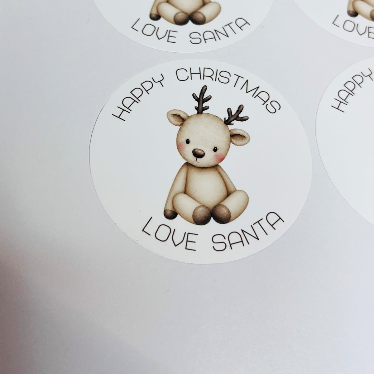 Round white Christmas stickers featuring a cute teddy bear reindeer with antlers and the text 'HAPPY CHRISTMAS LOVE SANTA' on a white background.