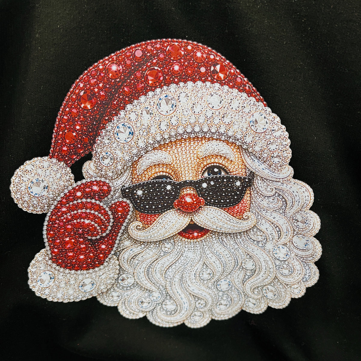 A close-up of a rhinestone-embellished Santa Claus face patch or applique on a black fabric background. The design features a red hat and mittens, a white beard and trim, black sunglasses, and a red nose, all covered with sparkling rhinestones.